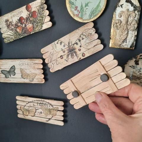 A hand holding popsicle sticks that are attached together and have various designs, including butterflies and plants. On of the pieces is facing away from the camera and has magnets adhered to the back.
