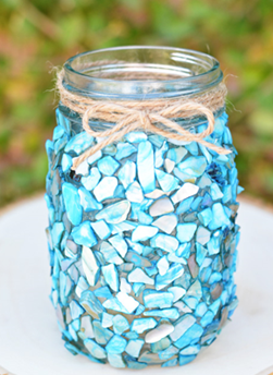 A decorated jar covered with blue and turquoise mosaic pieces, tied with a twine bow at the neck, displayed on a wooden slice.