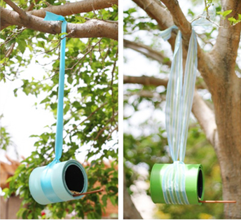 Two empty paint cans hanging from trees. The one on the left is painted blue and hanging from a blue ribbon and the one on the right is green hanging from a green ribbon.