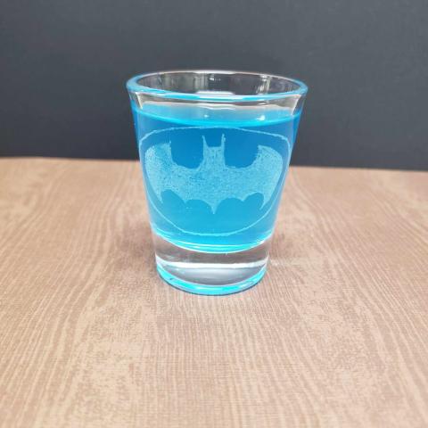 A photo of a shot glass with the Batman emblem engraved on it. It is full of a blue liquid and is sitting on a wooden surface.
