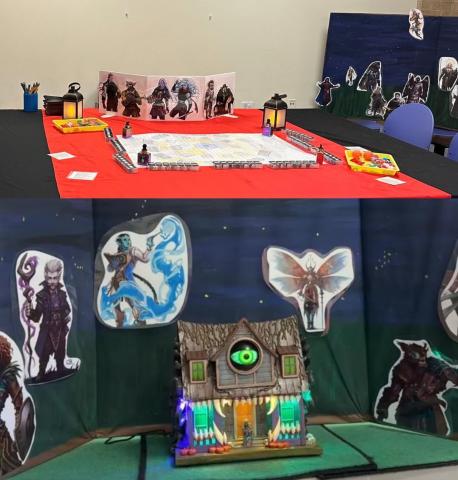 A table is covered in red and black table clothes has a map, fidgets, and characters on it for Dungeons and Dragons. Below is a Mimic House with a glowing eye and teeth against a night sky background surrounded by fantasy character portraits. 