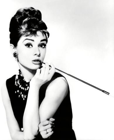 Black and white photo of Audrey Hepburn in an updo and black dress, holding a cigarette holder