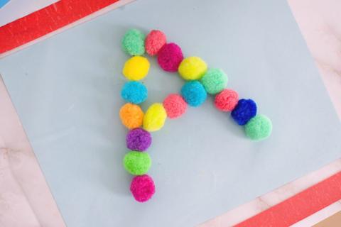 A large capital letter A is made out of many colored pom poms on a piece of paper