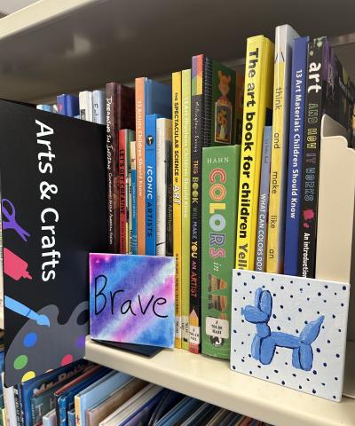 Two small paintings on a library shelf. The shelf is filled with art books. The left painting has blue, pink, and purple stripes and says "BRAVE" the right painting is a blue balloon dog with a blue polkadot background. 