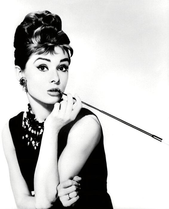 Black and white photo of Audrey Hepburn in an updo and black dress, holding a cigarette holder
